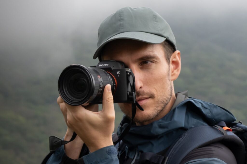 A photographer outdoors holding a Sony Alpha camera to his eye, framing a shot in a misty mountain environment.