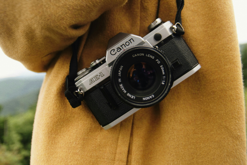 Canon AE-1 35mm film SLR with FD 50mm f/1.8 lens hanging against a tan wool coat outdoors — one of the cameras that were ugly when they launched but became icons