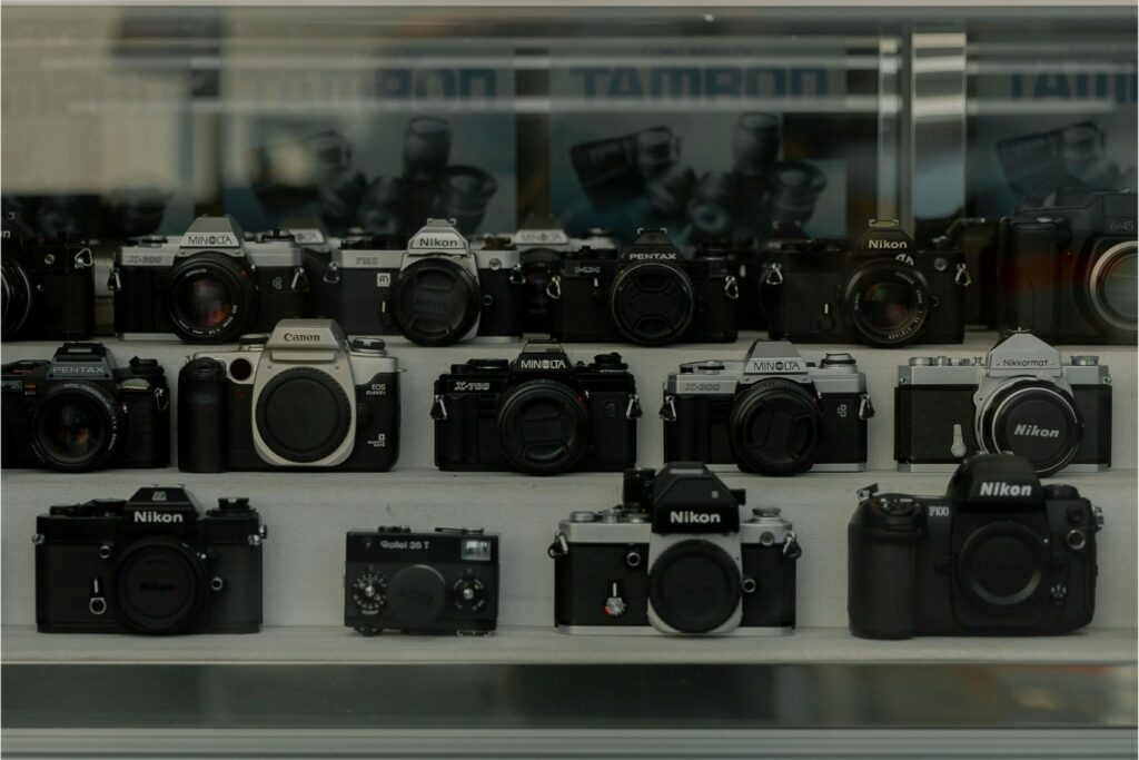 Collection of used cameras including Nikon, Canon, Minolta, and Pentax film bodies displayed on shelves behind glass