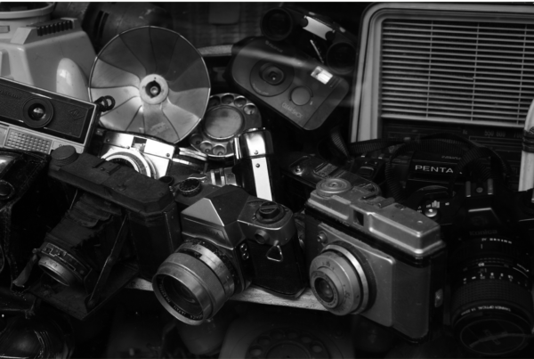 Camera Companies Make Cameras Nobody Wants — pile of discarded vintage film cameras, flash bulbs, and obsolete gear.