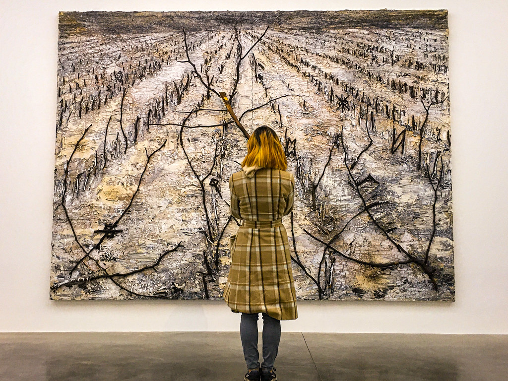 person in a plaid coat viewing a large landscape artwork on a white gallery wall