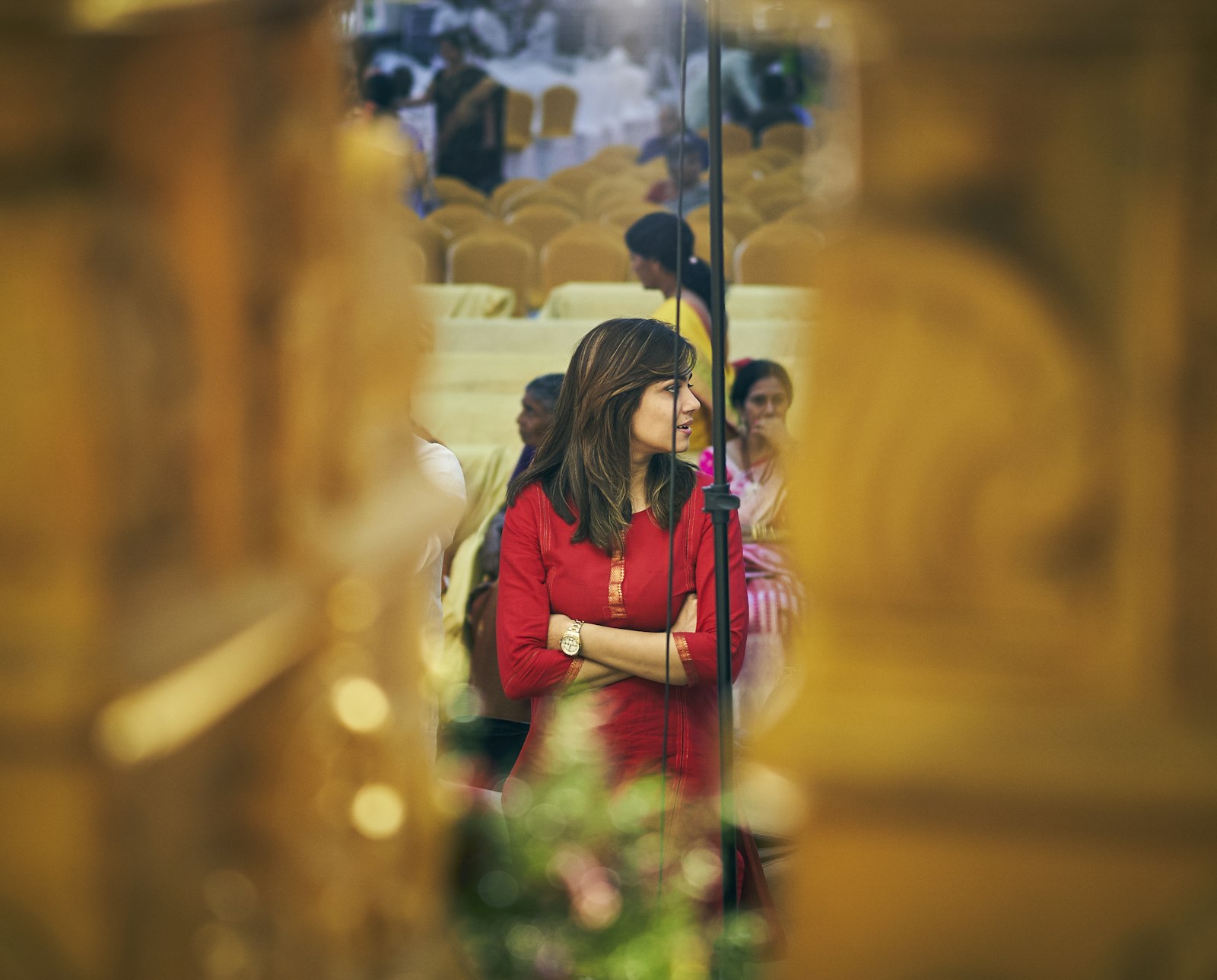 woman in red dress glancing aside at a warmly lit reception, seen through blurred foreground elements