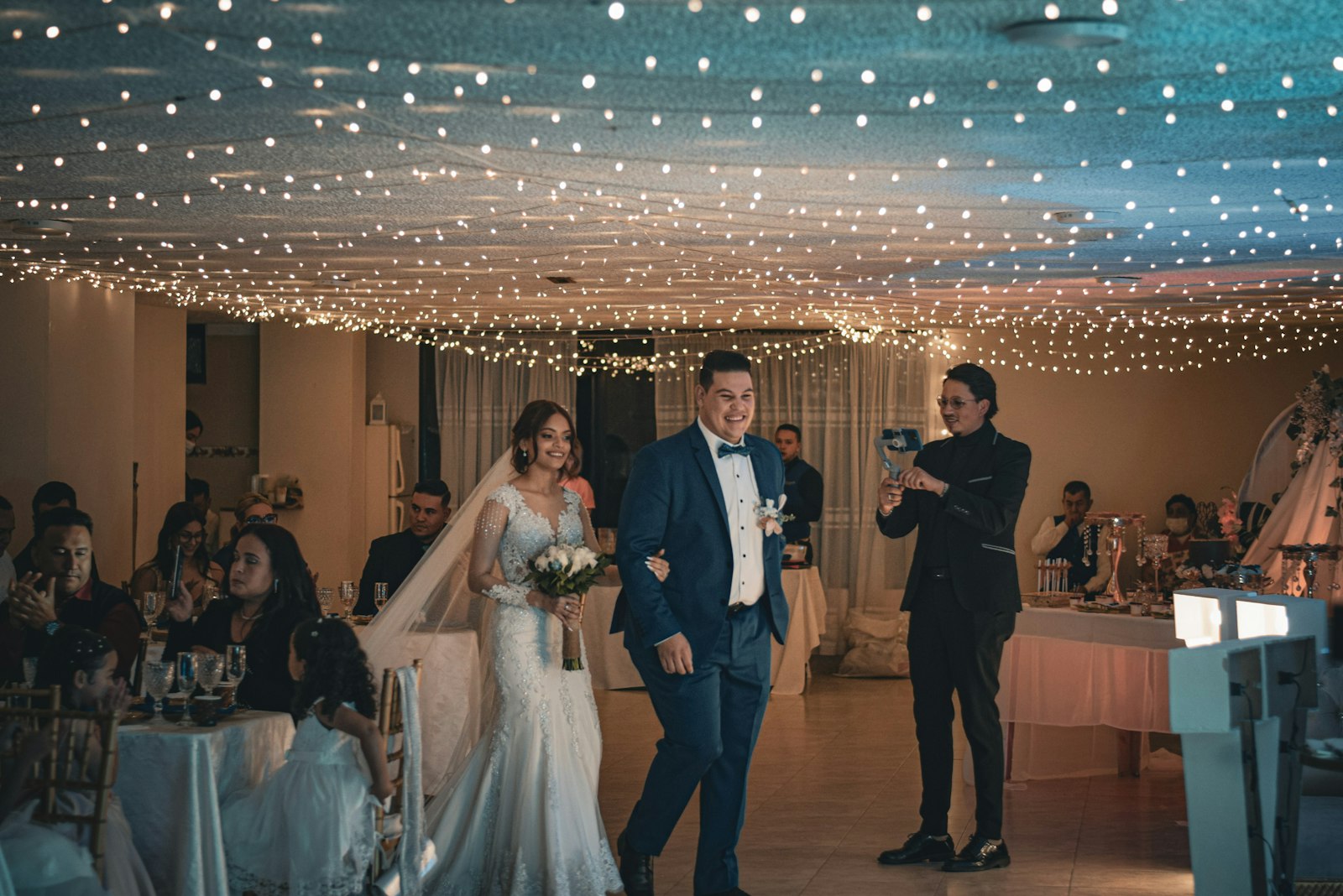 bride and groom walking through a wedding reception under string lights with guests seated at tables