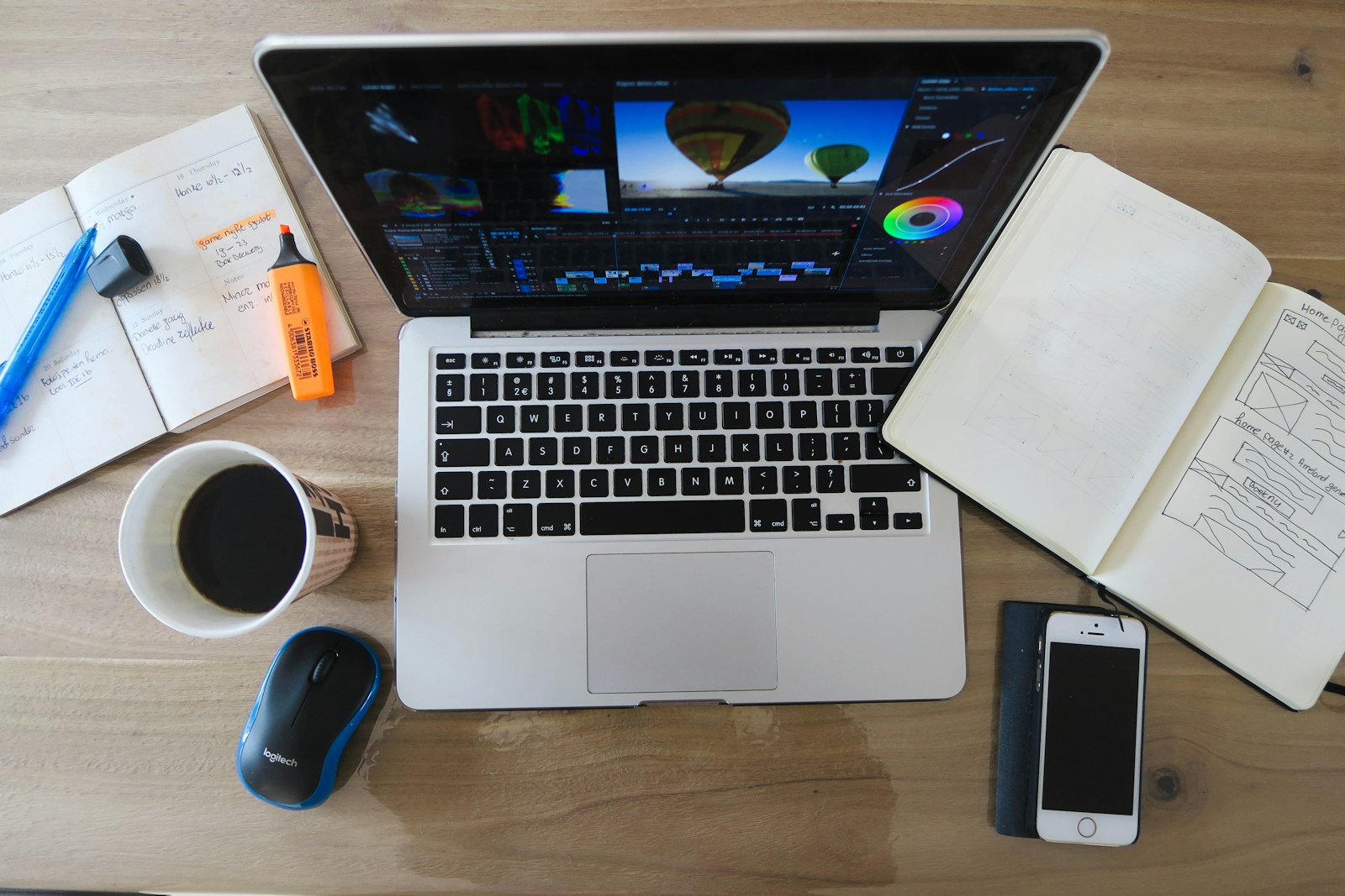 laptop with photo editing software beside a smartphone, notebooks, and coffee on a wooden desk