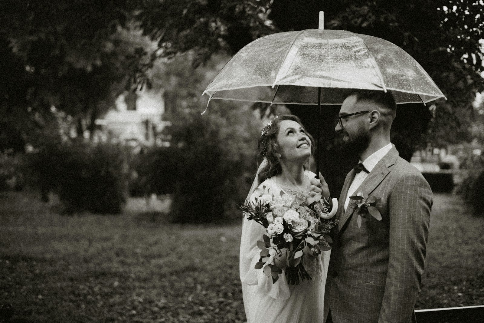 bride and groom standing under a clear umbrella outdoors surrounded by greenery