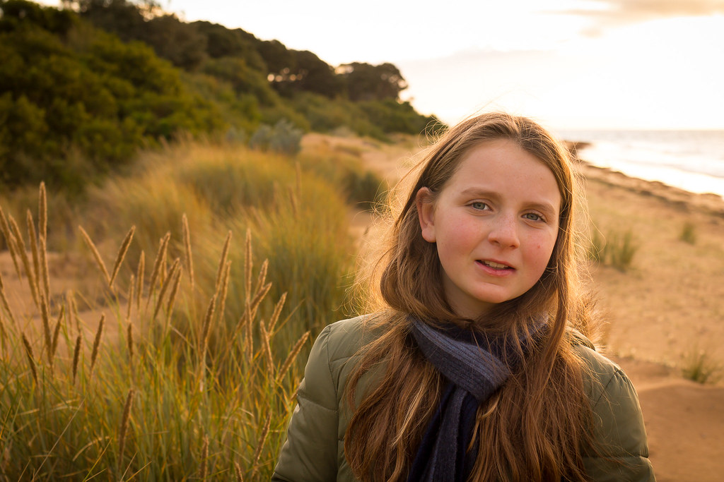 young woman with long hair in a green jacket photographed outdoors on a beach at sunset with blurred background