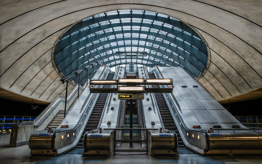 wide-angle view of an underground station with escalators beneath a domed glass ceiling