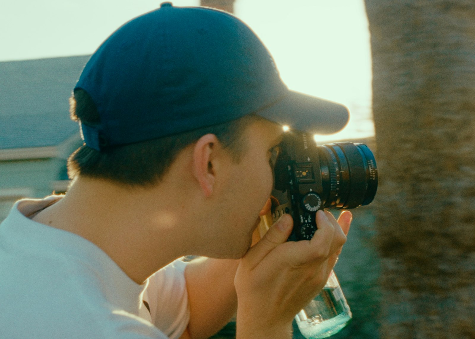 man in a cap and white shirt holding a black DSLR camera to his eye outdoors in warm light