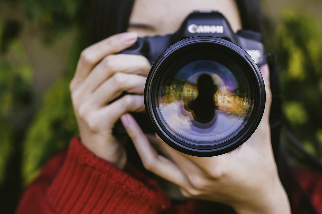 person holding a Canon DSLR camera with large lens facing the viewer, greenery in background