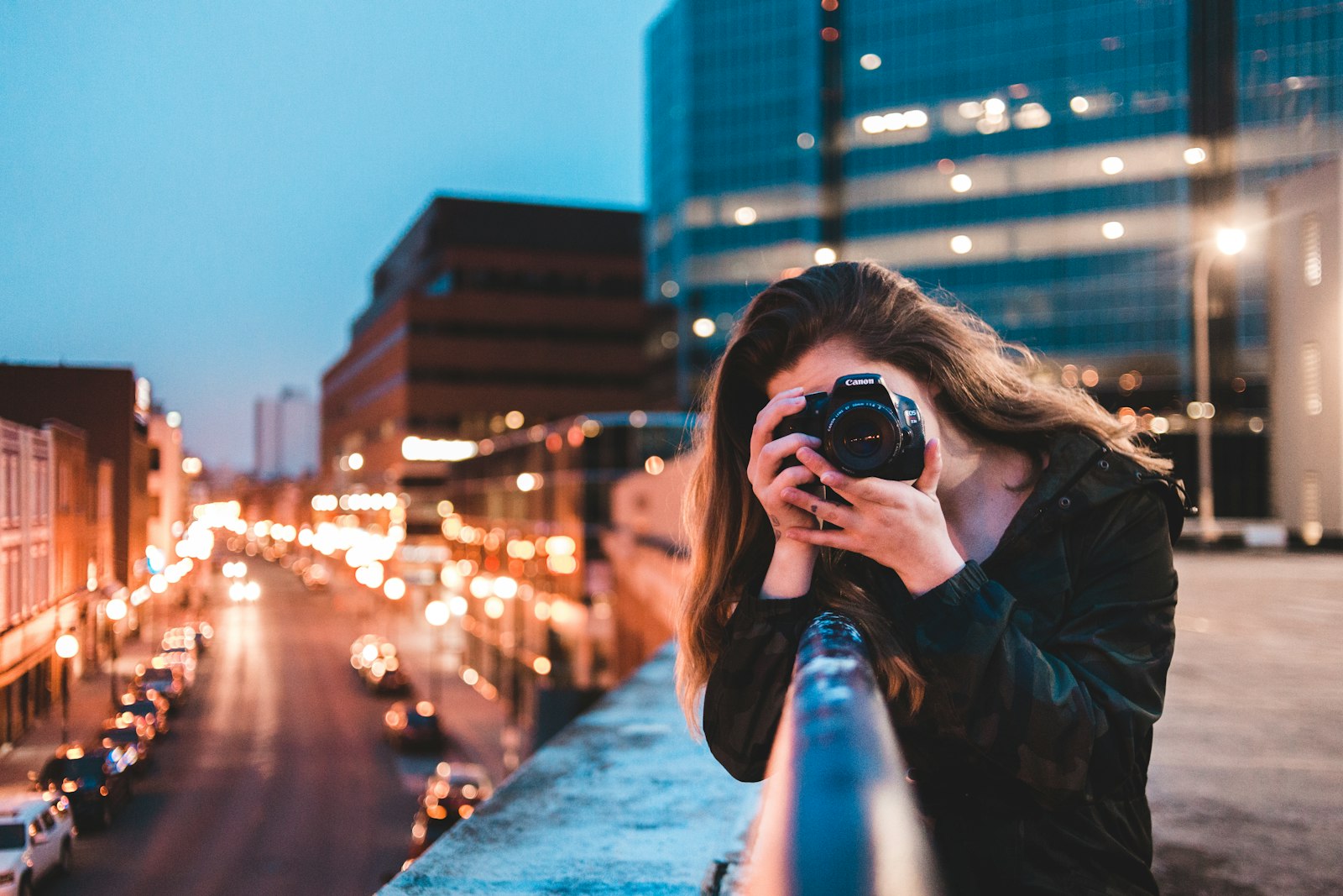 photographer holding a black camera to their eye with a city skyline and street lights behind at dusk