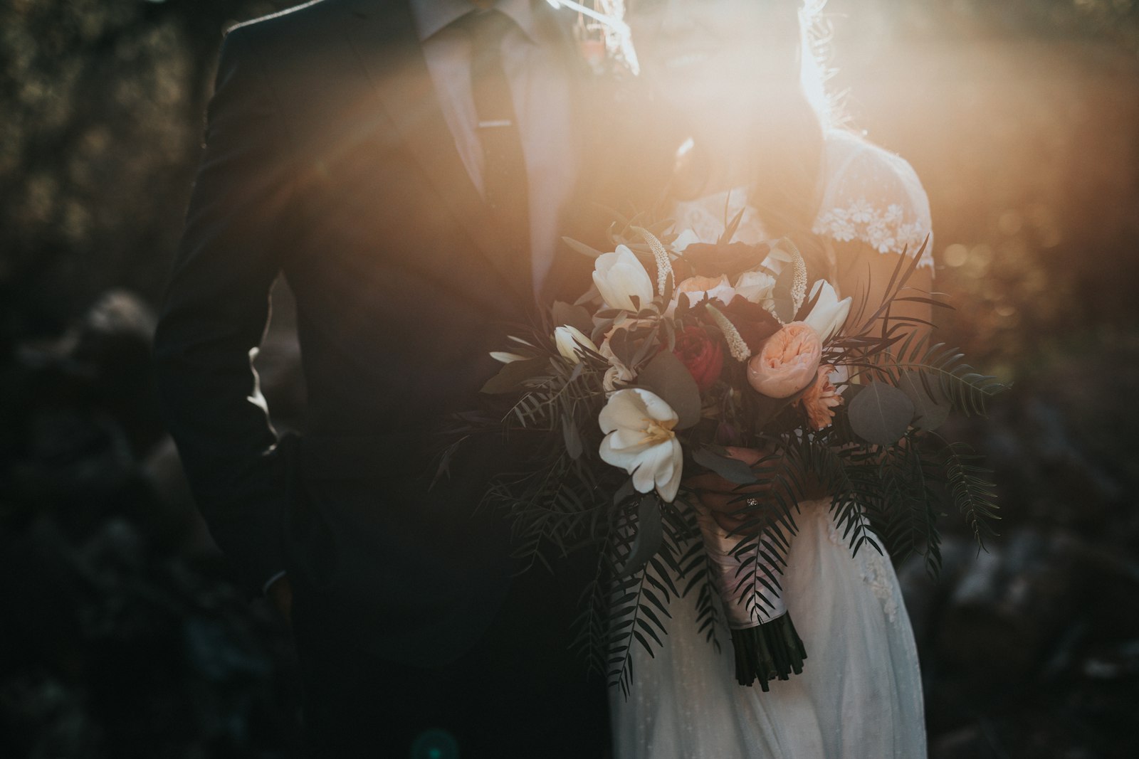 groom in dark suit beside bride holding a bouquet backlit by warm light