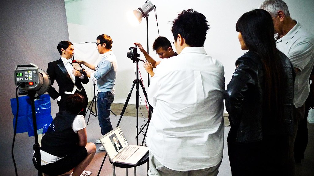 photography crew shooting a subject in a studio with strobes, a tripod-mounted camera, and a tethered laptop