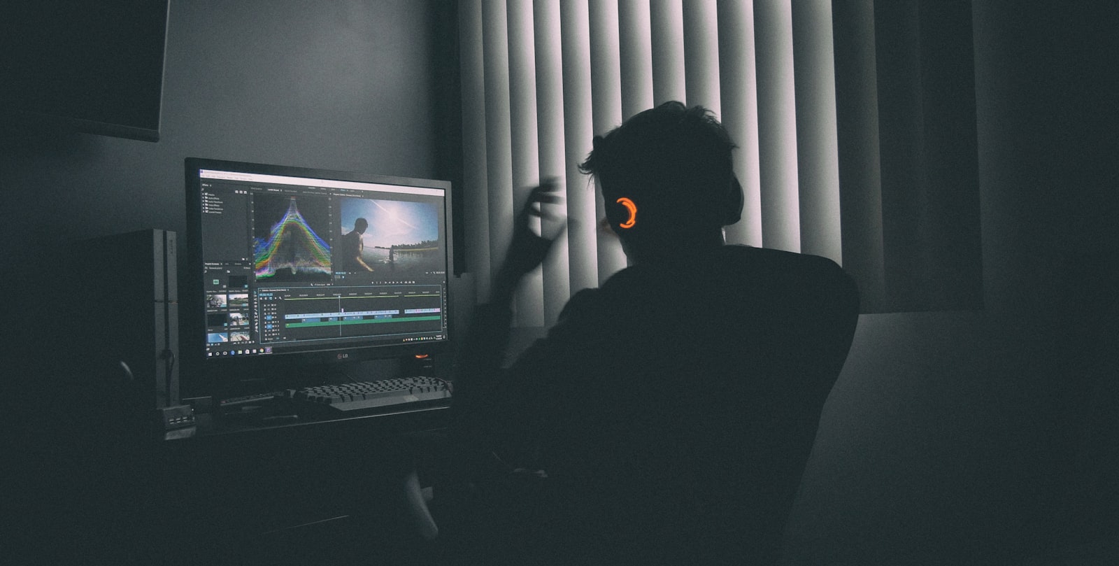 person in silhouette wearing headphones editing video footage on a computer in a dark room