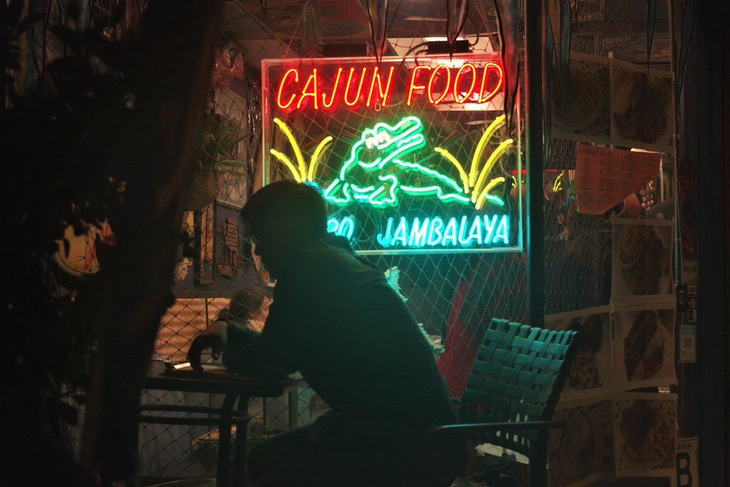 silhouette of a person seated at a table in front of a glowing Cajun Food neon sign at night