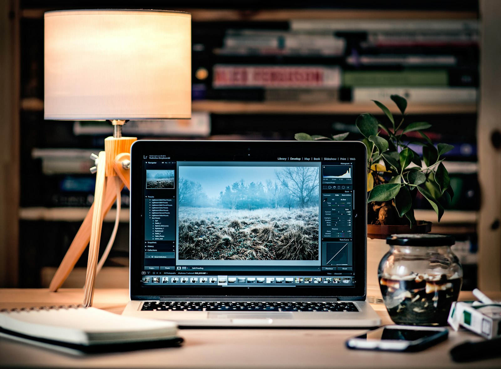 laptop displaying photo editing software on a desk with a lit lamp and plant in a dim room
