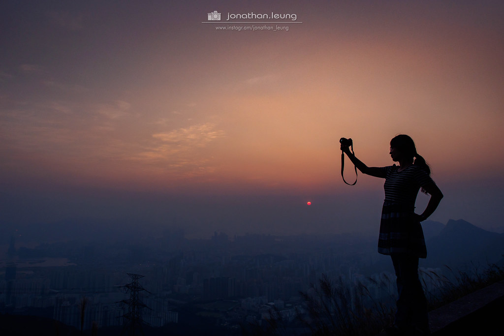 silhouette of a person holding a camera against a hazy sunset over a city skyline