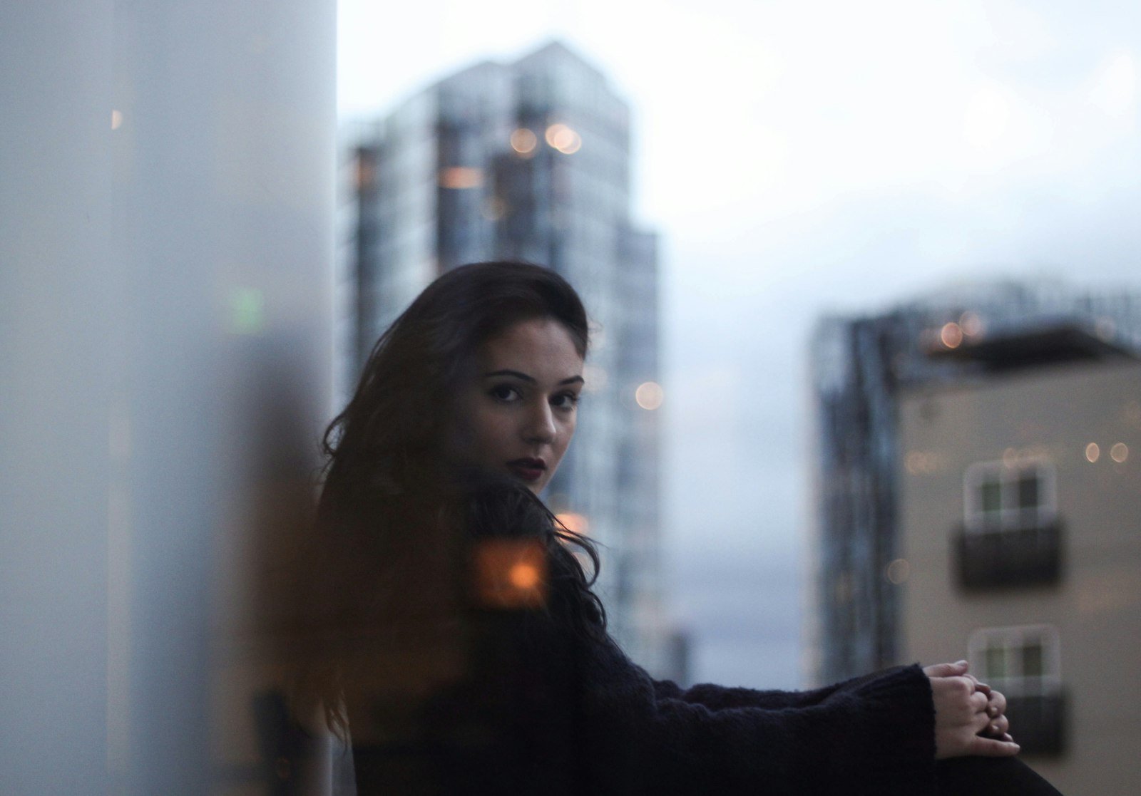 woman leaning against a window frame with a blurred city skyline behind her at dusk