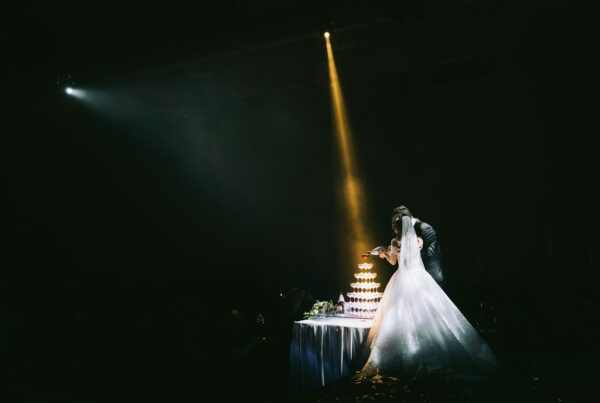bride and groom beside a tiered cake under a single spotlight in a dark reception hall