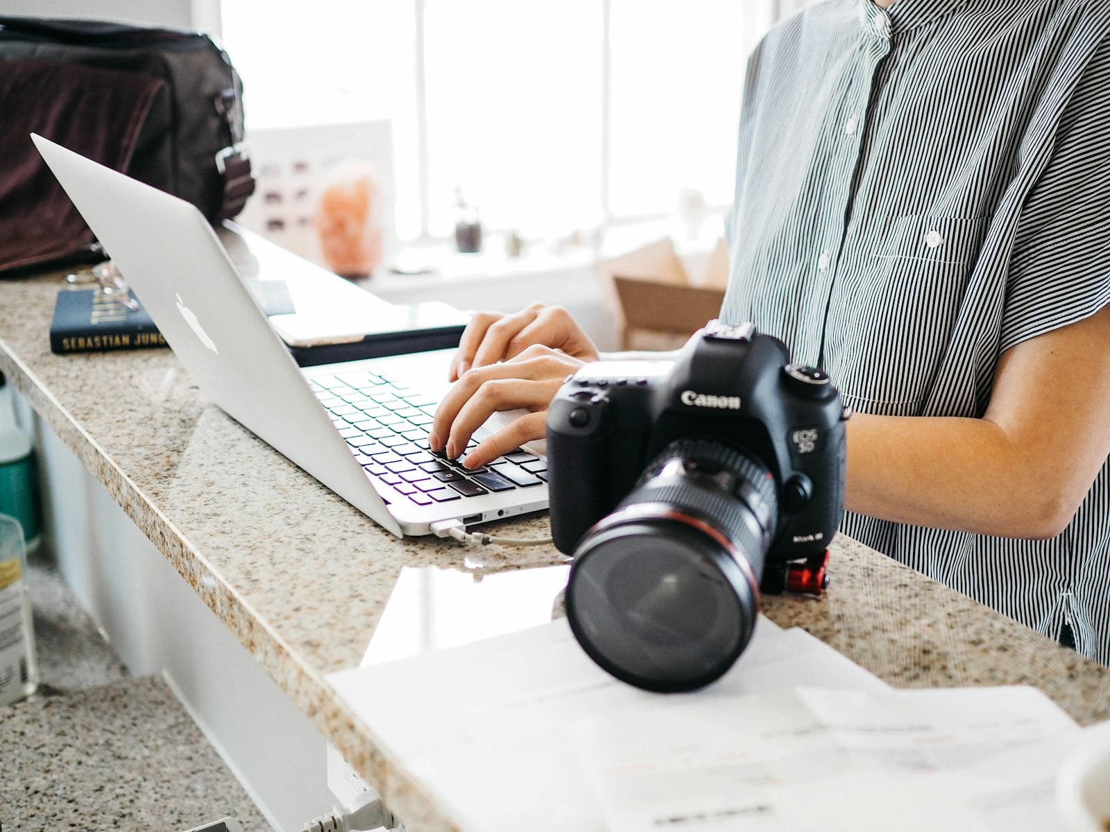 person working on a laptop at a desk with a DSLR camera and lens in the foreground