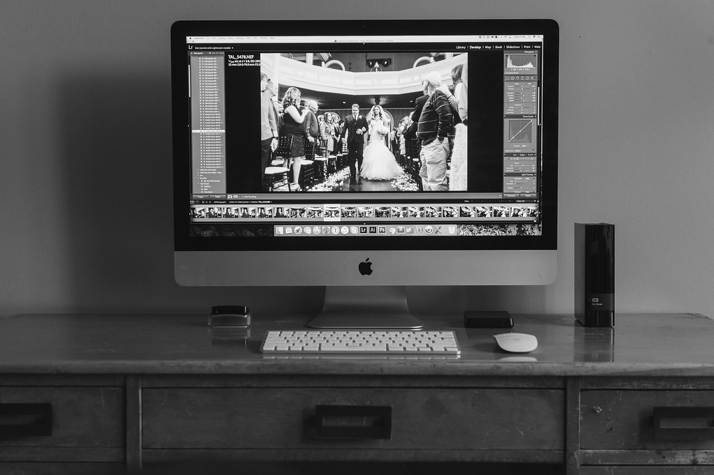 iMac displaying photo editing software with a wedding photo, keyboard, mouse, and external hard drive on a wooden desk