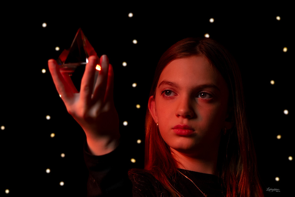 young person holding a glass pyramid, lit warmly from one side against a dark background with bokeh lights