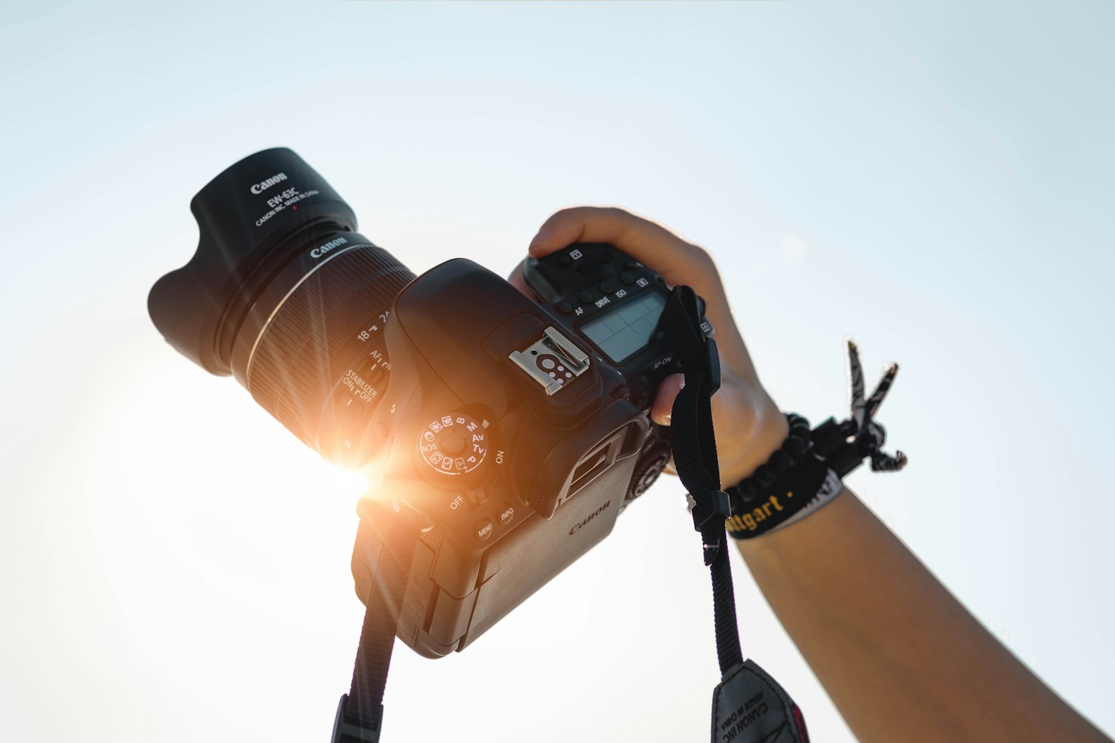 hands holding a Canon DSLR camera up against a bright sky with sun flare