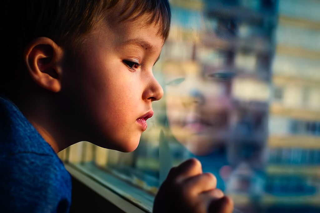 young boy lit by soft window light gazing out a window indoors